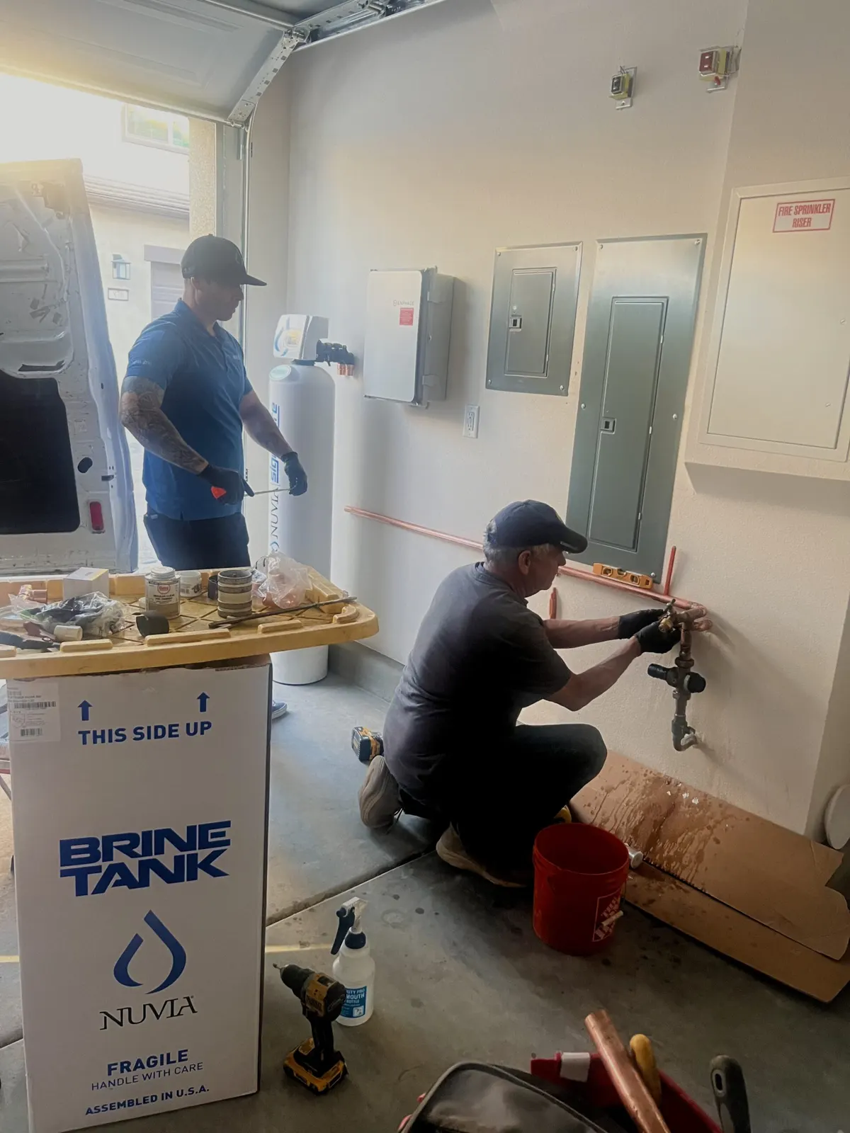 Two Nuvia installers fitting a copper water line in a garage, with a Nuvia brine tank box in the foreground