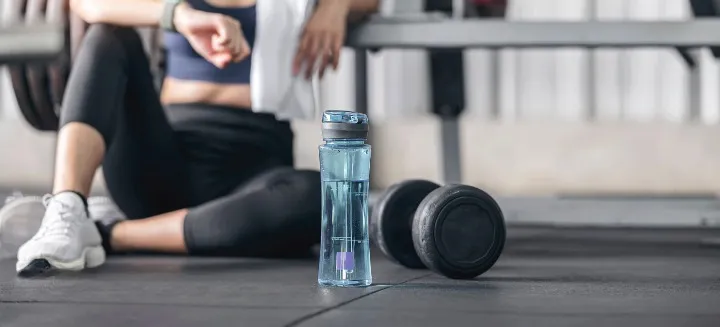 A filled water bottle on a gym floor next to a pair of dumbbells